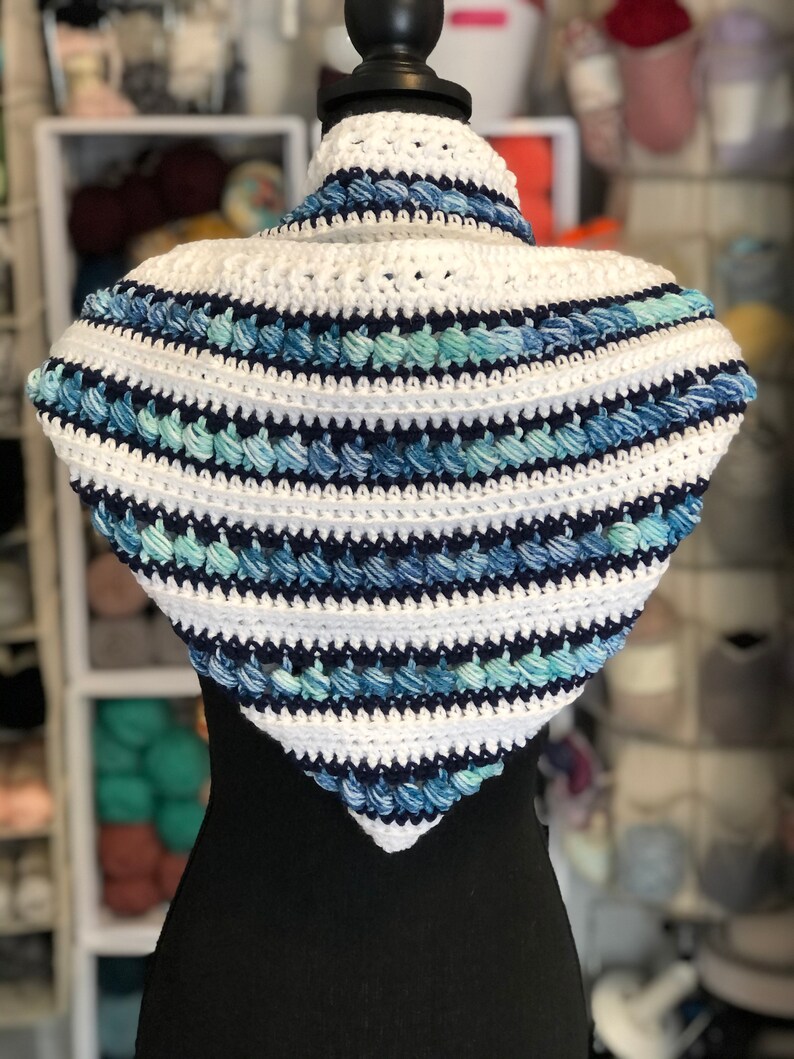May include: A white and blue crocheted triangular scarf with a textured design. The scarf is displayed on a black mannequin.