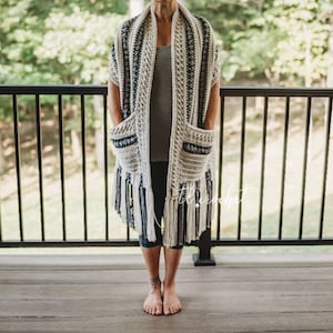 Op de afbeelding: Een wit en zwart gehaakt sjaal met franjes en twee zakken. De sjaal wordt gedragen door een persoon die op een houten dek staat.