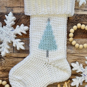 May include: White crocheted Christmas stocking with a blue and gray embroidered Christmas tree design. The stocking is on a wooden surface with wooden beads and snowflakes.