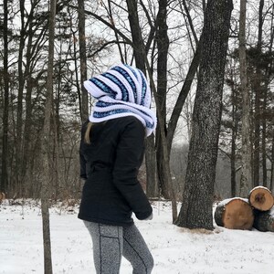May include: A white and blue striped crocheted hooded scarf with a long, flowing design. The scarf is being worn by a person in a snowy woodland setting.