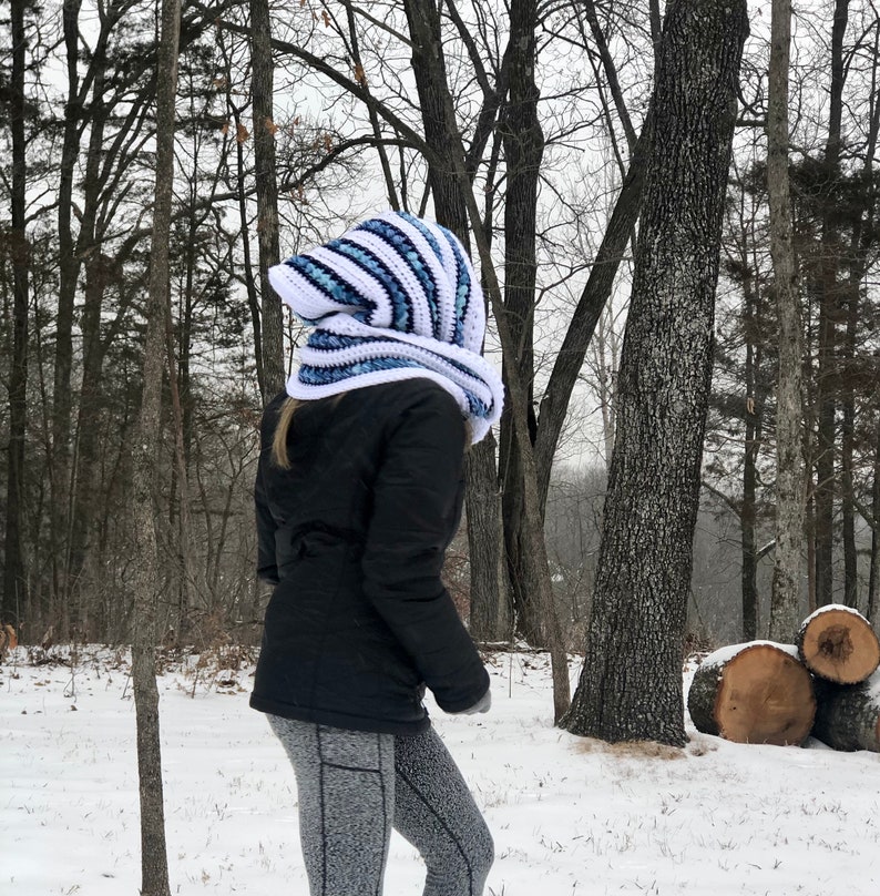 May include: A white and blue striped crocheted hooded scarf with a long, flowing design. The scarf is being worn by a person in a snowy woodland setting.