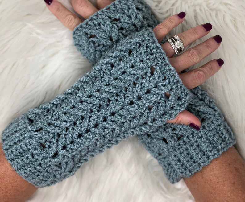 Puede incluir: Un par de guantes sin dedos de color azul hechos a crochet con un patr&oacute;n de encaje. Los guantes se llevan en las manos de una persona.
