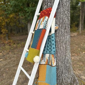 May include: A colorful, patchwork crochet scarf with pom-poms hangs on a white wooden ladder leaning against a tree. The scarf features various textured patterns and a palette of teal, orange, yellow, and rust colors. The background shows a blurred forest scene.