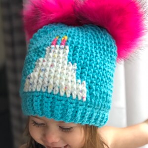 May include: A turquoise blue knitted hat with a white unicorn design and two pink pom poms on top. A young child is wearing the hat.