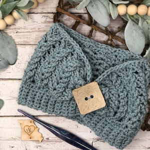 Puede incluir: Un paño de cocina de crochet azul verdoso con un botón cuadrado y un gancho de crochet de madera con una etiqueta en forma de corazón que dice "TLC".