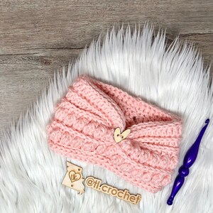 Puede incluir: Una diadema de crochet rosa con un botón de corazón de madera y el texto "@tl.crochet" en una etiqueta de madera.