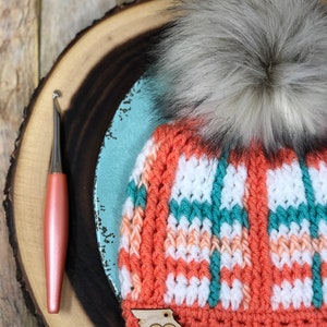 May include: A crocheted beanie hat with a white, peach and teal plaid pattern. The hat has a fluffy white pom pom and a wooden tag with the letters "TLC" and a heart.