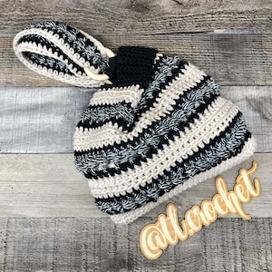 Op de afbeelding: Een zwart-wit gestreepte gehaakte tote bag met een houten handvat. De tas heeft een textuur en is gemaakt van dik garen. De tas ligt op een houten oppervlak. De tekst "@ttl.crochet" staat op een houten bord op de voorgrond.
