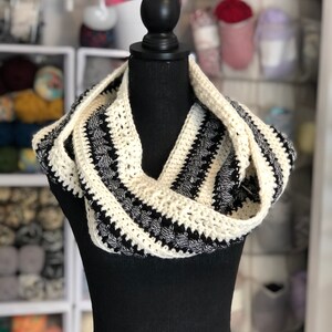May include: A black and white crocheted infinity scarf with a textured design. The scarf is displayed on a black mannequin.