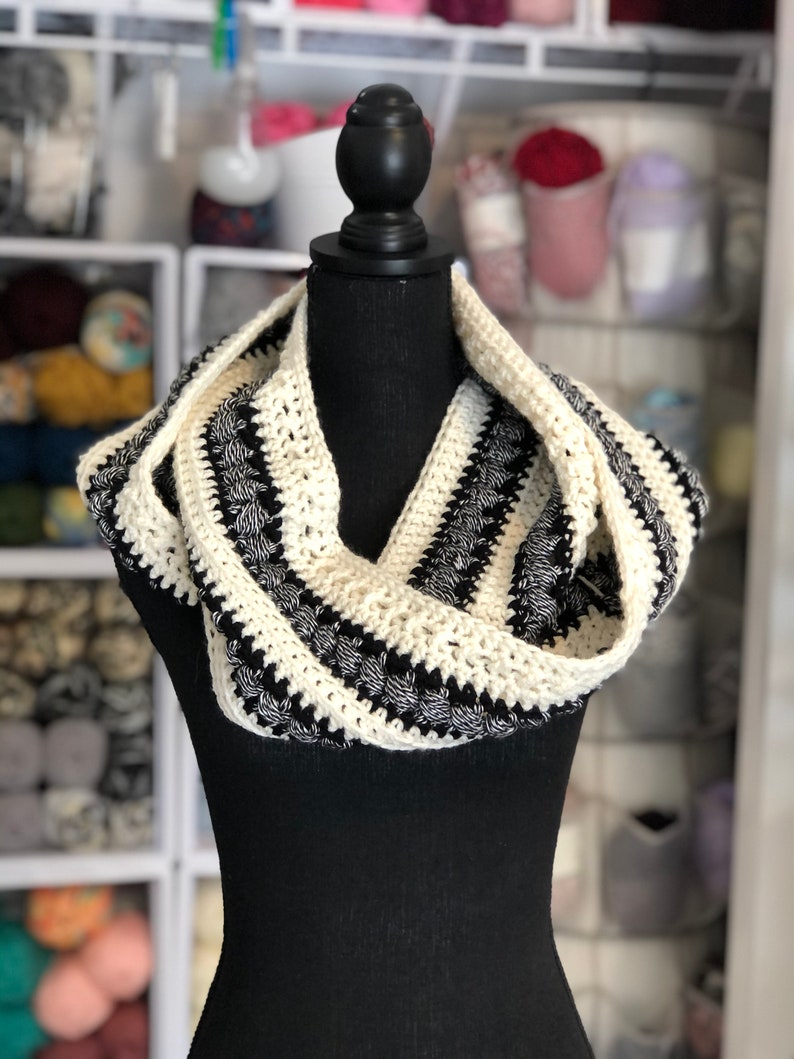 May include: A black and white crocheted infinity scarf with a textured design. The scarf is displayed on a black mannequin.