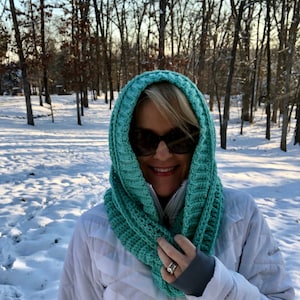 May include: A woman wearing a white jacket and a teal blue crocheted cowl neck scarf. The scarf is worn over her head and covers her ears. She is smiling and looking at the camera. The background is a snowy forest.