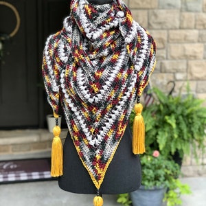 Op de afbeelding: Een driehoekige gehaakte sjaal met een geometrisch patroon in grijstinten, zwart, rood en geel. De sjaal heeft een franje rand en kralenkwastjes.