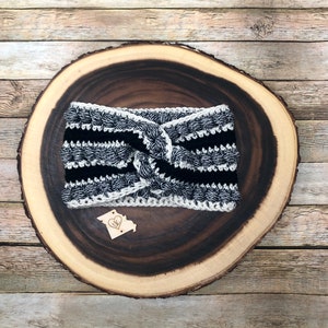 Op de afbeelding: Zwart-wit gestreept gehaakt hoofdband met een geknoopt ontwerp. Het hoofdband wordt getoond op een houten oppervlak.