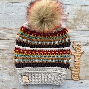 Puede incluir: Un gorro de ganchillo hecho a mano en una variedad de colores, incluyendo marrón, rojo, amarillo y azul. El gorro presenta un gran pompón de piel sintética en la parte superior. Una pequeña etiqueta de madera con un contorno en forma de corazón de Missouri está adherida al borde. El texto "tl crochet" es visible.