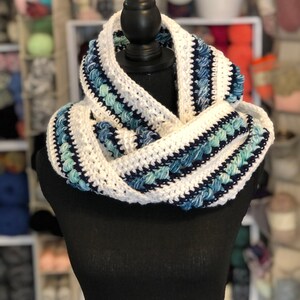 May include: White and blue crocheted infinity scarf with a textured design. The scarf is displayed on a black mannequin.