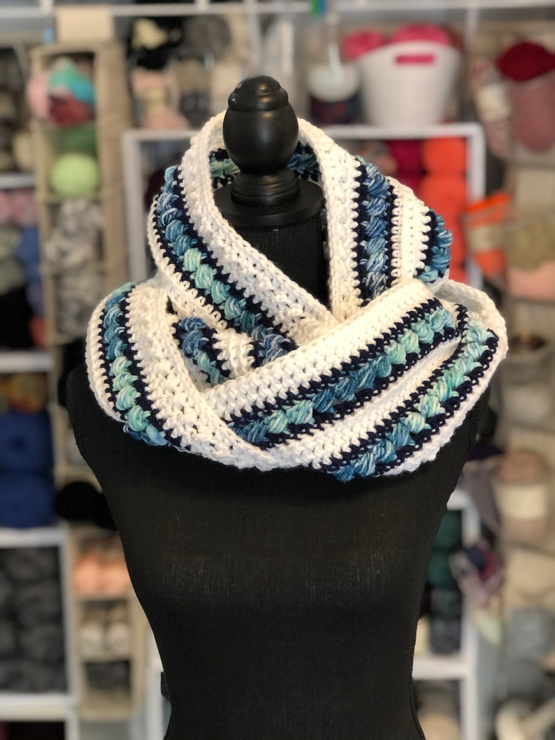May include: White and blue crocheted infinity scarf with a textured design. The scarf is displayed on a black mannequin.