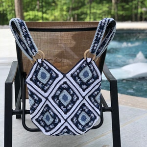 Op de afbeelding: Een gehaakte tote bag met blauwe en witte geometrische patronen en houten handvatten. De tas hangt aan een zwarte stoel bij een zwembad.