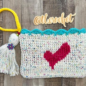 May include: A hand-crocheted clutch bag with a white speckled pattern, a bright pink heart, and a turquoise scalloped edge. It features a yellow wrist strap and a white tassel with a flower accent. The text "@tl.crochet" is visible.