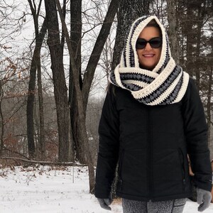 May include: A woman wearing a black hooded jacket and a white and black striped crocheted infinity scarf with a hood. The scarf is worn over her head and covers her ears. She is standing in a snowy woodland.