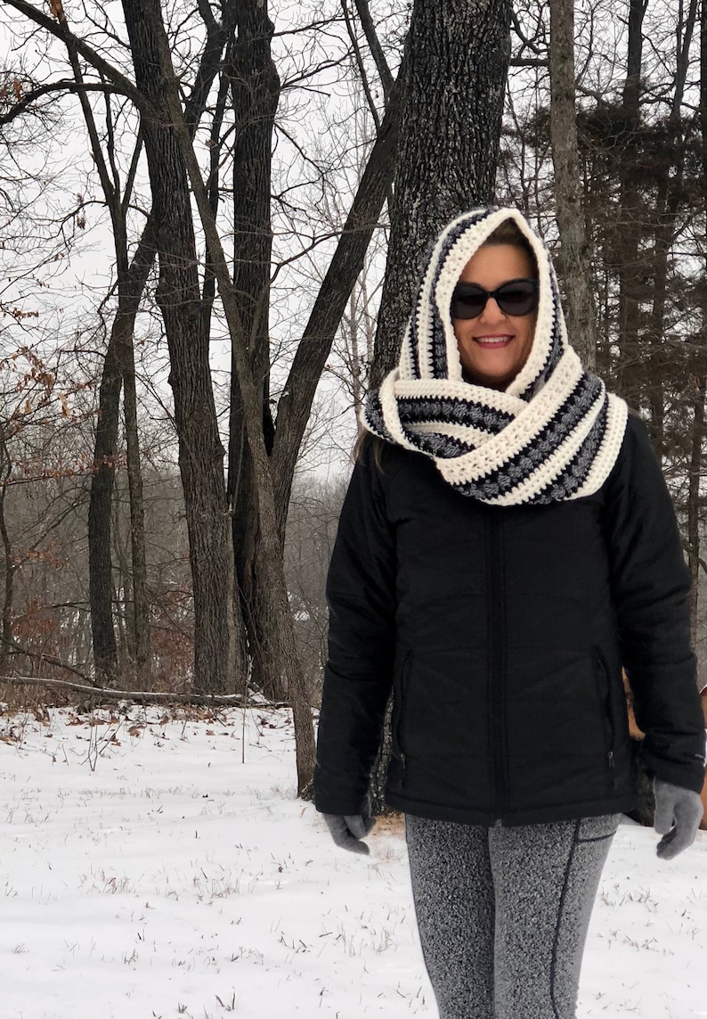 May include: A woman wearing a black hooded jacket and a white and black striped crocheted infinity scarf with a hood. The scarf is worn over her head and covers her ears. She is standing in a snowy woodland.