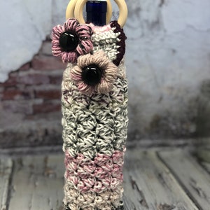 Puede incluir: Una funda de botella de vino de ganchillo con un esquema de color rosa, blanco y gris. La funda tiene un diseño floral decorativo con botones negros. La funda está coronada con un asa de anillo de madera.