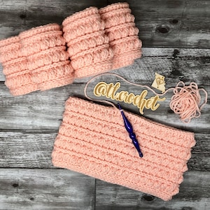 May include: A close-up of a pink crocheted headband with a textured stitch pattern. The headband is on a wooden surface with a wooden tag that says "@ttl.crochet".