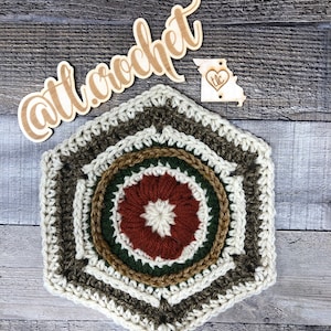 Puede incluir: Un hexágono de ganchillo con un centro multicolor en tonos marrones, verdes y blancos. El centro tiene un diseño de flor. El hexágono está rodeado por un borde blanco y un borde marrón y verde.