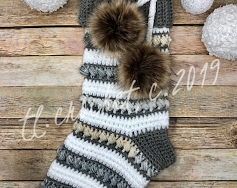 May include: A gray and white striped crocheted Christmas stocking with two brown faux fur pom poms. The stocking is hanging on a wooden background with white decorative balls.