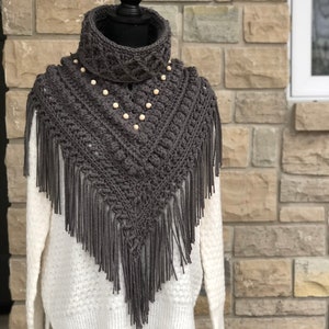 Op de afbeelding: Grijze gehaakte sjaal met franjes en houten kralen. De sjaal is op een witte trui op een mannequin.