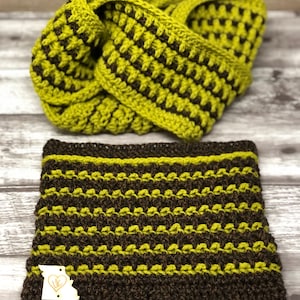 May include: A brown and green crocheted infinity scarf and matching headband. The scarf is folded in half and the headband is lying on top of it. The headband has a small tag with the text "LLC" on it.