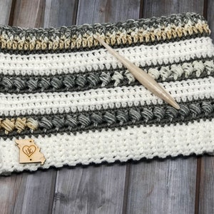 Op de afbeelding: Een gehaakte sjaal in wit, grijs en beige met een houten label waarop "MO" in een hart staat. De sjaal ligt op een houten oppervlak met een haaknaald.