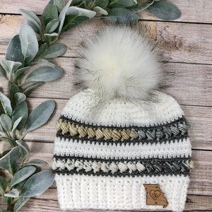 Op de afbeelding: Witte gehaakte beanie met een nep bont pompon, bruine en grijze strepen en een houten patch met een hart en de letters "MO".
