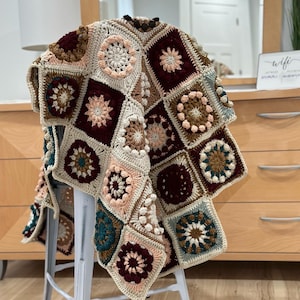 Puede incluir: Una manta de ganchillo con un diseño de patchwork que presenta cuadrados en tonos de marrón, beige, rosa, verde azulado y burdeos. La manta está drapeada sobre un taburete blanco.