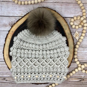May include: A crocheted beanie hat with a faux fur pom pom in a light gray color. The hat has a textured design with small beads around the brim.