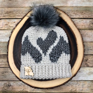 Puede incluir: Un gorro de punto gris y negro con un pompón de piel sintética negra. El gorro tiene un diseño en forma de corazón con un patrón blanco y negro. El gorro está sobre una superficie de madera. El gorro tiene una pequeña etiqueta de madera adjunta con las letras "ILC" y un corazón.