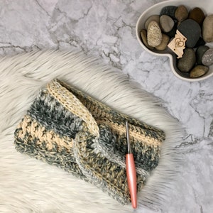 Op de afbeelding: Een gehaakt hoofdband in beige en grijze tinten met een roze haaknaald op een wit nep bont oppervlak.