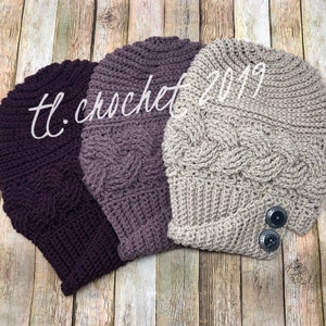 Puede incluir: Tres gorros de ganchillo en diferentes tonos de morado y beige. Los gorros tienen un patrón de punto de cable y presentan un detalle de botón.
