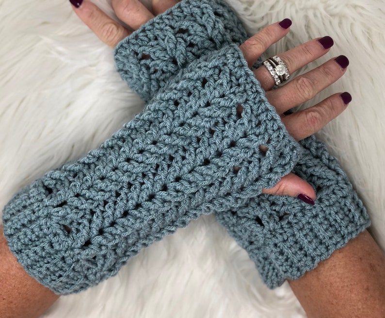 Puede incluir: Un par de guantes sin dedos de color azul hechos a crochet con un patr&oacute;n de encaje. Los guantes se llevan en las manos de una persona.