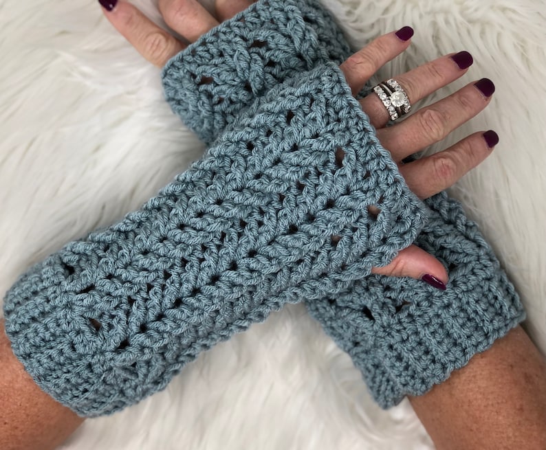 Puede incluir: Un par de guantes sin dedos de color azul hechos a crochet con un patr&oacute;n de encaje. Los guantes se llevan en las manos de una persona.