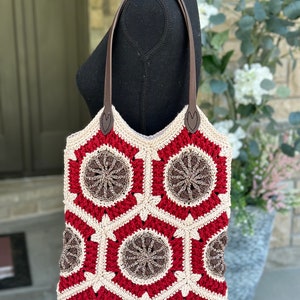May include: A crocheted tote bag with a geometric pattern in red, brown, and cream. The bag has brown leather handles.