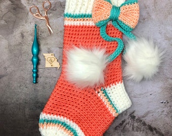 May include: A crocheted Christmas stocking in coral, white, and teal with a large white pom-pom and a smaller white pom-pom. The stocking has a large bow made of coral and teal yarn.