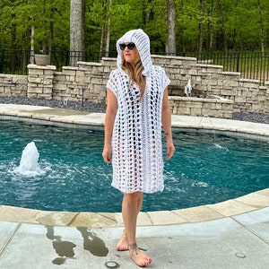 May include: A white crochet cover-up with a hood, worn by a person standing by a swimming pool.
