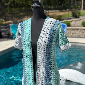 May include: A crocheted cardigan with a V-neck and short sleeves. The cardigan is made in a striped pattern of light blue, teal, and gray yarn.
