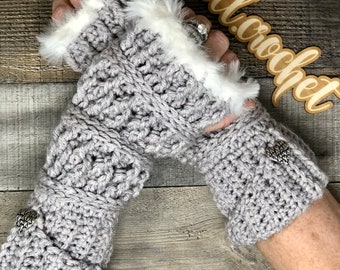 Puede incluir: Guantes sin dedos de crochet grises con un puño de piel sintética blanca y adornos en forma de corazón plateados. Los guantes se muestran sobre un fondo de madera con el texto "@tt.crochet" en un letrero de madera.