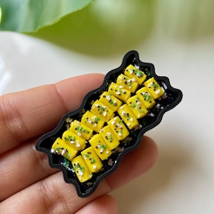 Puede incluir: Miniatura de sushi en un plato negro. El sushi amarillo está cubierto con semillas negras y hierbas verdes. El plato se sostiene en una mano, con una hoja verde en el fondo.