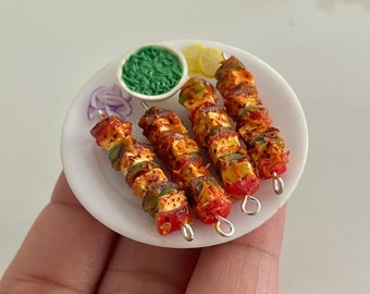 May include: Four miniature skewers of paneer tikka on a white plate with a small bowl of green chutney and a lemon wedge. The skewers are made of polymer clay and are decorated with red, green, and white details.