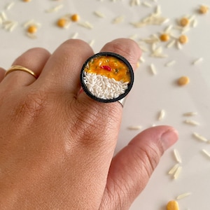 Puede incluir: Un anillo con una banda negra y un diseño de comida redondo, blanco y naranja. El diseño de la comida parece un tazón de arroz con salsa de curry encima.