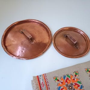 Puede incluir: Dos tapas de olla de cobre con asas. La tapa más grande es ligeramente más grande que la más pequeña.