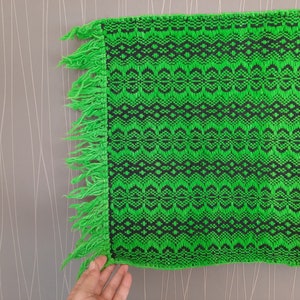 May include: Green and black woven fabric with a fringe border. The fabric has a geometric pattern.