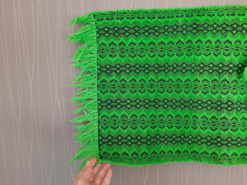 May include: Green and black woven fabric with a fringe border. The fabric has a geometric pattern.